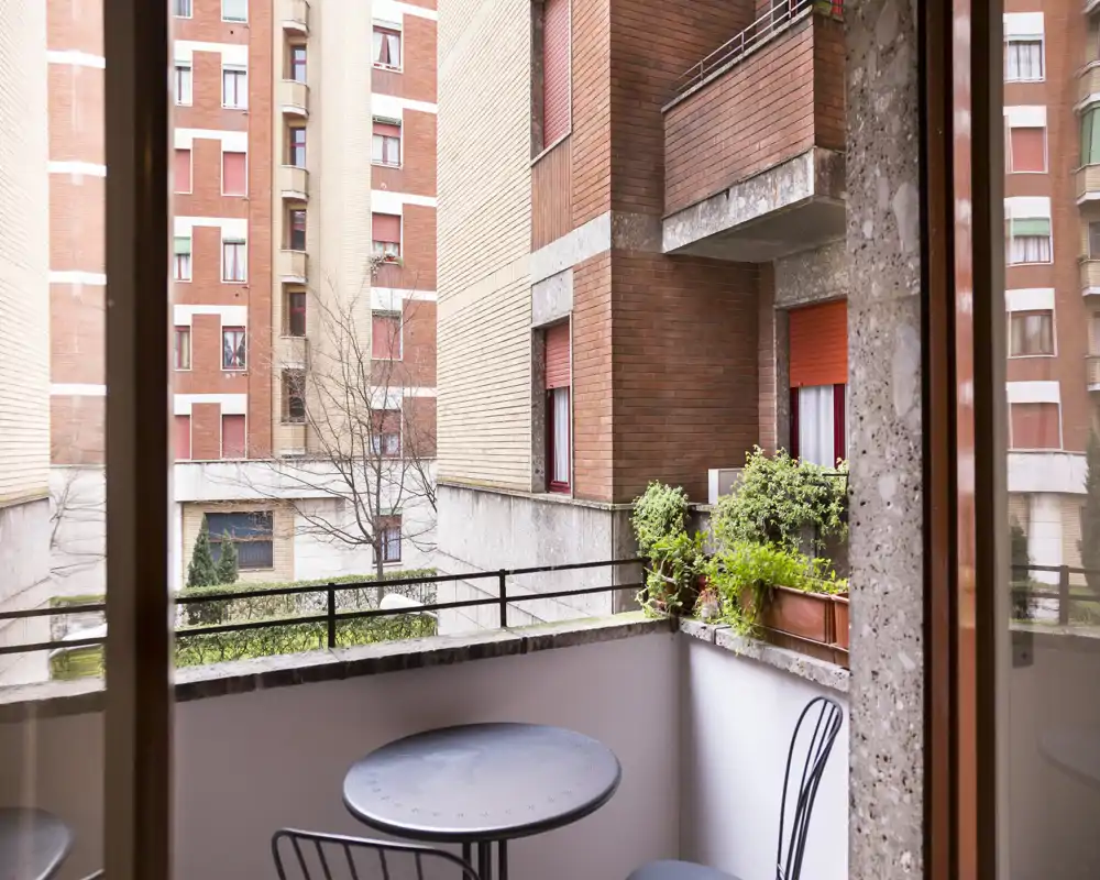 Pequeño balcón urbano con mesa redonda de metal, dos sillas y macetas, con vistas a edificios de ladrillo — espacio exterior luminoso y privado.