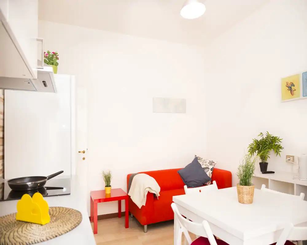 Zona de estar luminosa y moderna con un sofá rojo llamativo, mesa de comedor blanca y plantas decorativas — acogedora y bien iluminada, perfecta para relajarse y comer.
