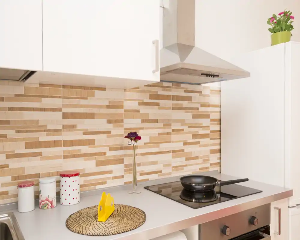 Rincón de cocina luminoso y compacto con vitrocerámica, campana extractora de acero y tarros decorativos sobre la encimera; salpicadero de azulejos efecto madera y un pequeño jarrón que aportan calidez.
