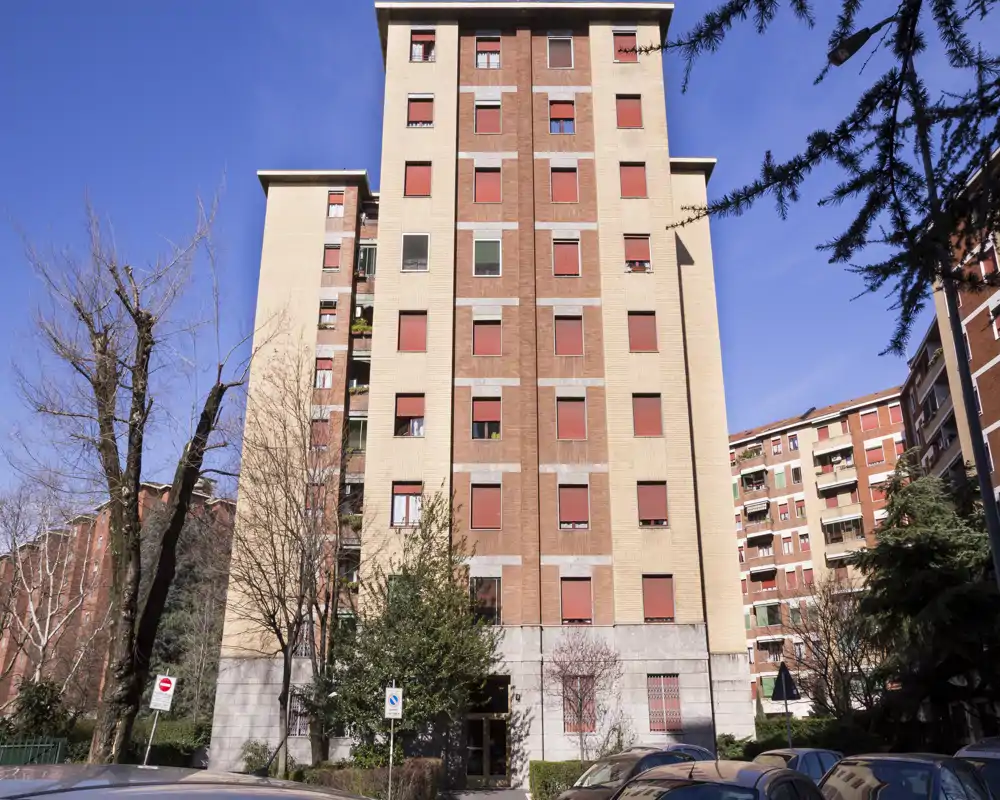 Vista exterior de un edificio residencial de varios pisos con persianas rojas y cielo azul, mostrando la fachada frontal y la entrada a nivel de calle.