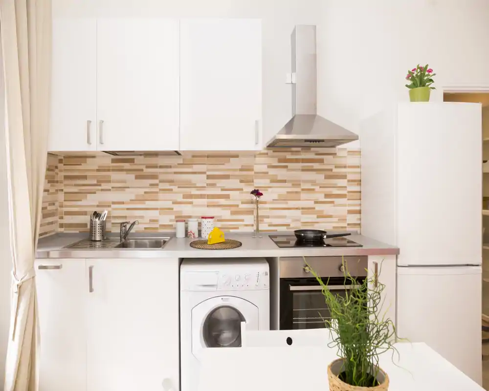 Cocina moderna y compacta, luminosa, con horno integrado, placa de cocción y lavadora, salpicadero de azulejos cálidos y muebles blancos.