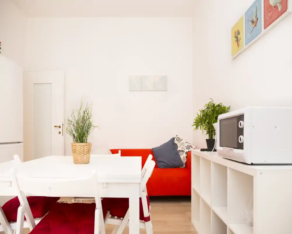 Área de estar luminosa y ordenada con sofá rojo, mesa de comedor blanca y estantería — bien iluminada y con mobiliario agradable, ideal para un apartamento compacto.