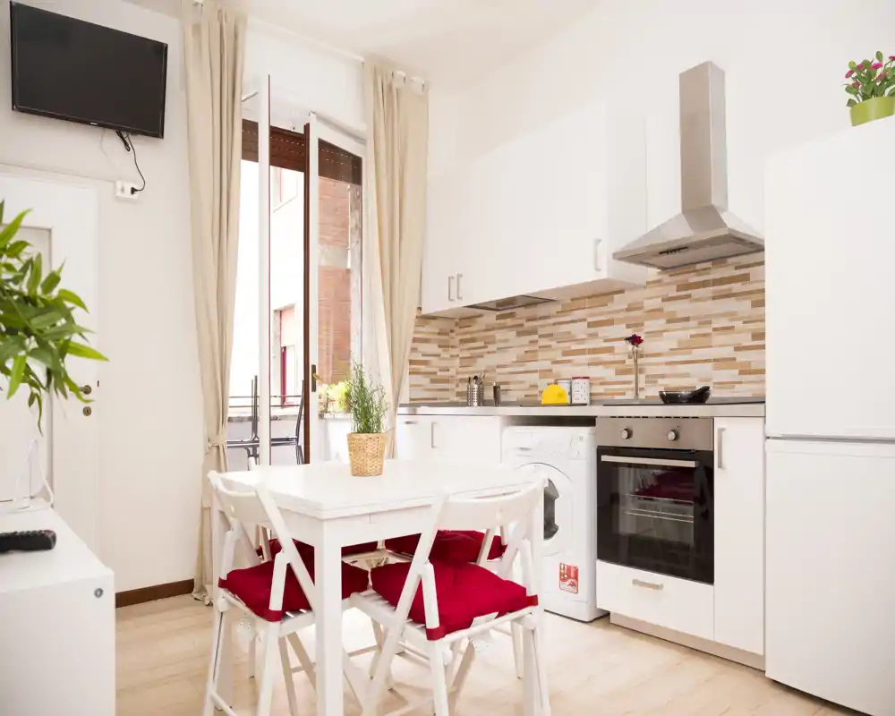 Cocina moderna y compacta con muebles blancos, horno empotrado, lavadora y una pequeña mesa con sillas con cojines rojos; luz natural desde la puerta abierta y un salpicadero de azulejos cálidos crean un ambiente agradable.