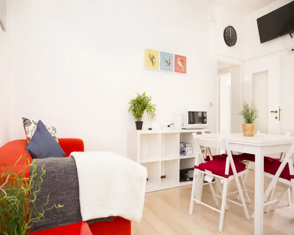 Sala luminosa y acogedora con sofá rojo, mesa de comedor blanca y sillas plegables, pequeño estante y plantas decorativas — ideal para un apartamento compacto.