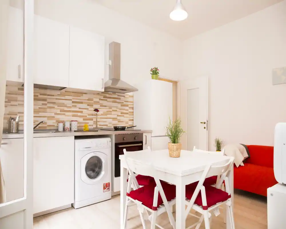 Cocina abierta y luminosa con muebles blancos, horno integrado, lavadora y una mesa de comedor pequeña con sillas con cojines rojos. El alicatado cálido y la planta en maceta crean un ambiente acogedor y bien decorado, perfecto para apartamentos compactos.