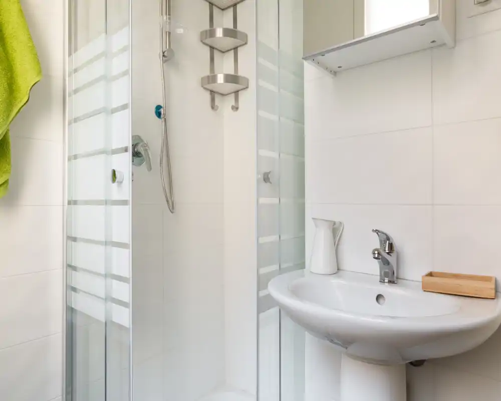 Baño moderno y luminoso con ducha en esquina, lavabo con pedestal y grifería cromada; azulejos blancos y accesorios sencillos.