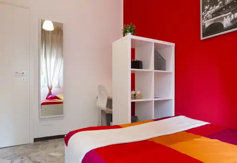 Dormitorio luminoso y colorido con pared roja, cama bien hecha con ropa de cama naranja y roja, una estantería blanca y un pequeño escritorio junto a la cama.