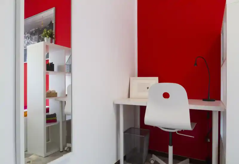 Pequeño espacio de trabajo con mesa y silla blancas frente a una pared roja intensa, con lámpara, cuadro y estantería ordenada — perfecto como rincón de oficina en casa.