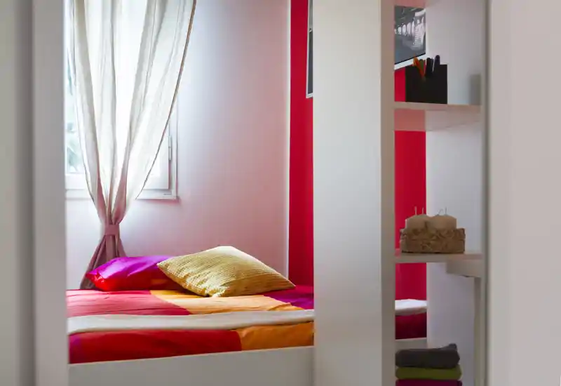 Dormitorio luminoso y colorido con cama arreglada, cojines decorativos y luz natural desde la ventana — cálido y acogedor.