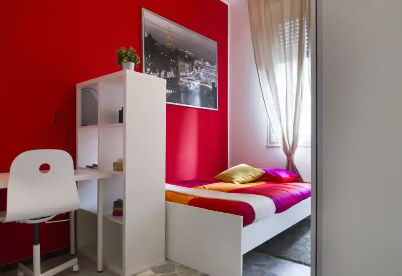 Dormitorio compacto y luminoso con una cama individual colorida, cojines suaves y un rincón de estudio ordenado. Una gran imagen en blanco y negro y cortinas translúcidas aportan estilo y dejan pasar la luz natural.