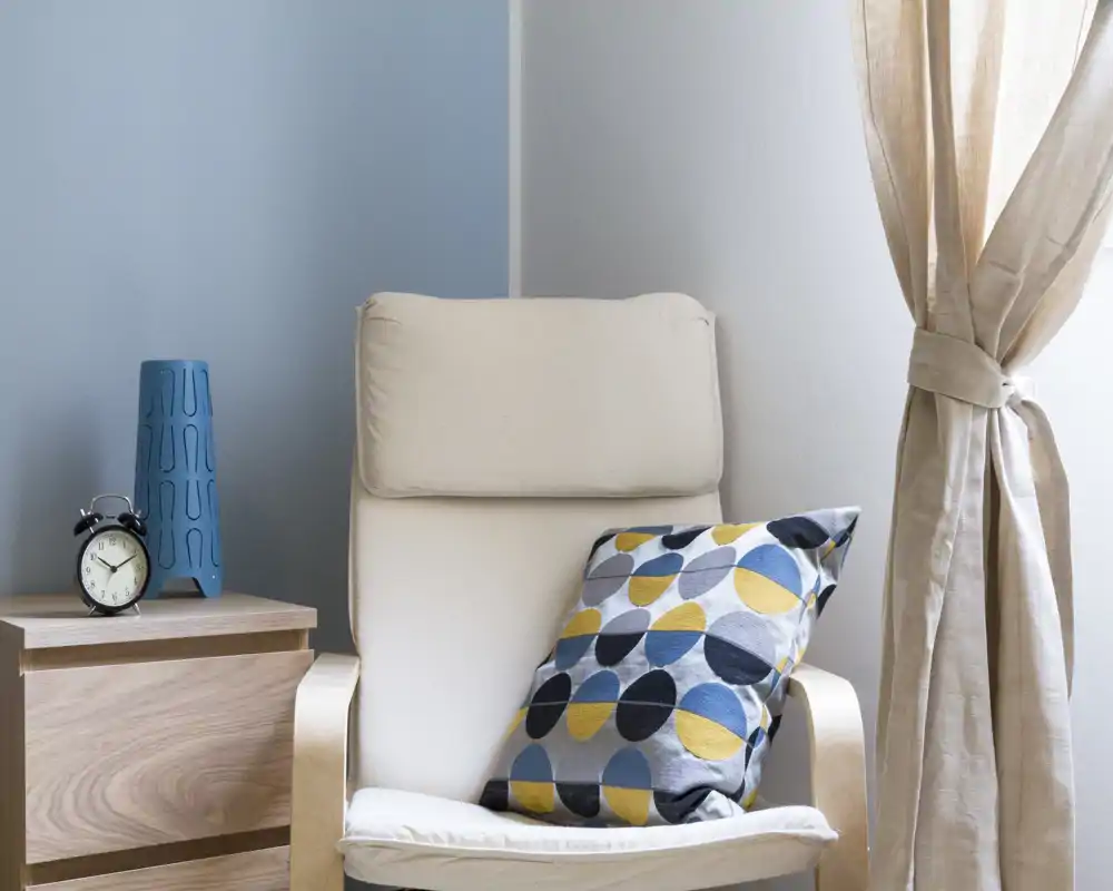 Cozy reading nook with a cushioned armchair, patterned throw pillow and soft natural light from a curtained window — ideal for quiet relaxation.