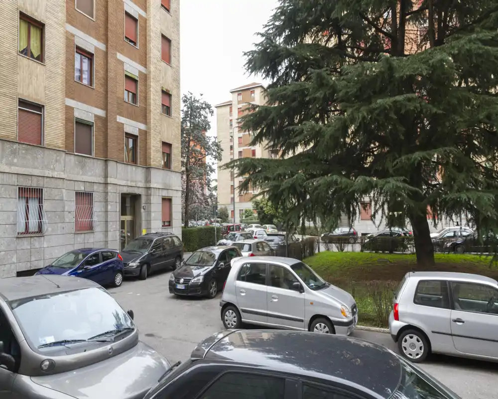 Communal courtyard/parking area in front of residential buildings with several parked cars and mature trees, well lit and clearly framed.
