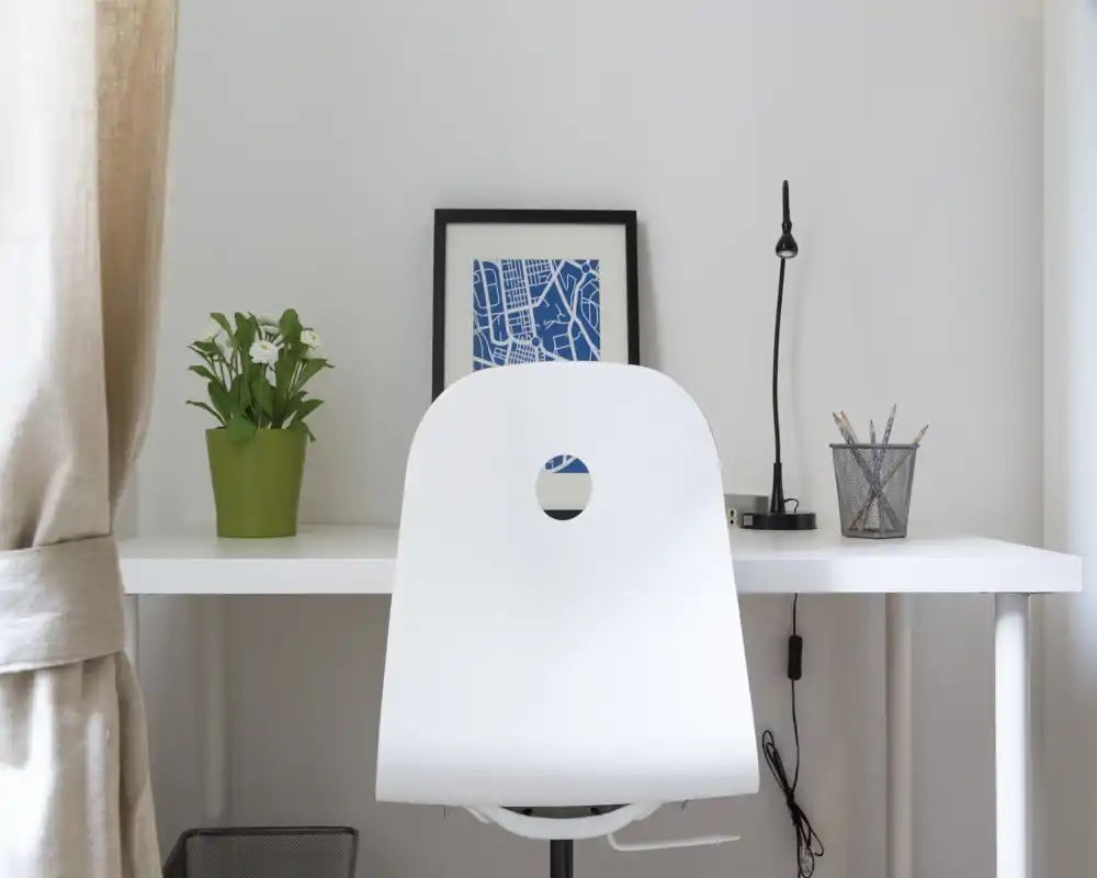 Bright, minimalist workspace with a white desk and chair, small lamp, potted plant and framed print — neat and well lit for remote work or study.