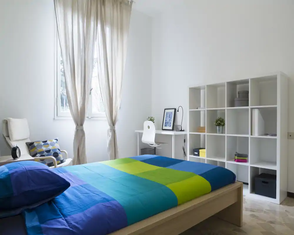 Bright, modern bedroom with a colorful blue-green duvet, a comfortable bed dominating the space, a desk by the window and open cube shelving for storage.