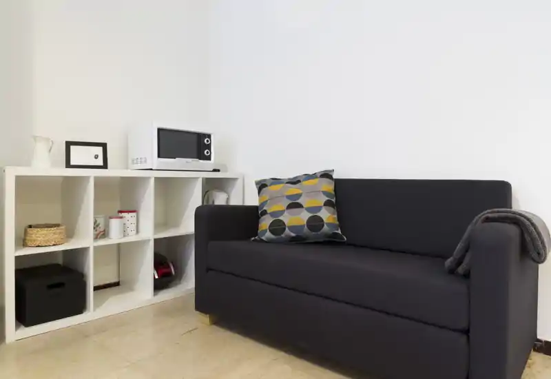 Compact living area with a dark upholstered sofa, patterned throw cushion and a ladder-style storage unit with decorative baskets and a microwave — bright, tidy and simply styled.