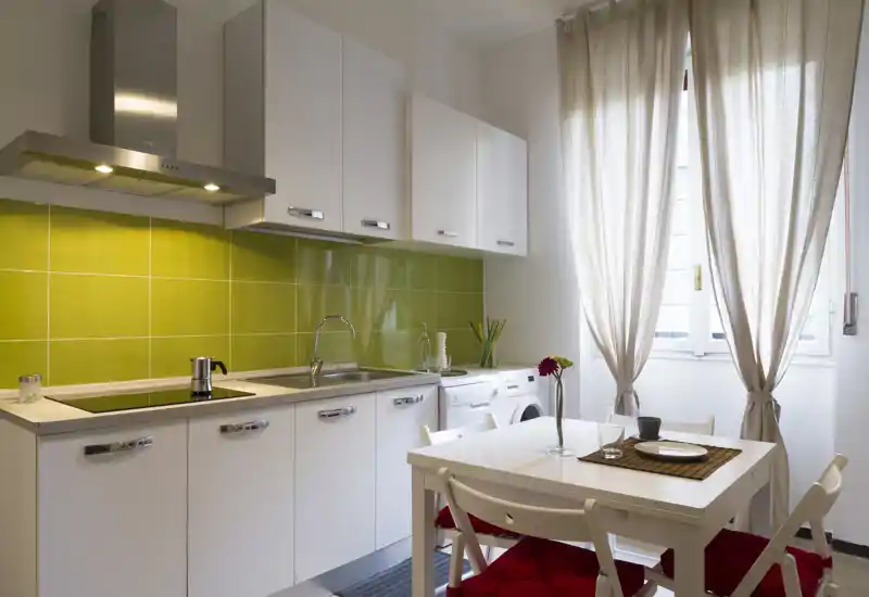 Bright, modern kitchen with white cabinets, a green tiled backsplash and a small dining table bathed in natural light — ideal for cozy meals.