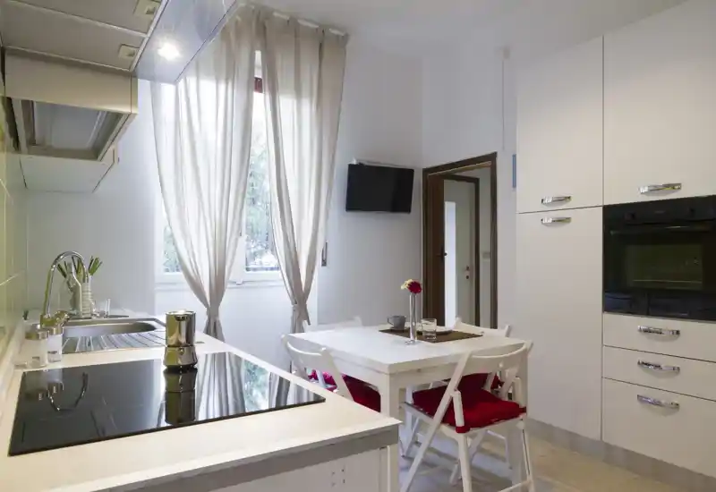 Modern compact kitchen with a built-in oven, induction cooktop and a small dining table. Light curtains and natural daylight create a bright, welcoming atmosphere.