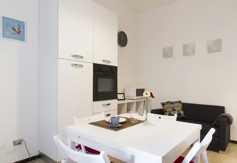 Bright, compact dining area with a white table and chairs, a built-in oven and cabinets, and a small sofa — ideal for studio or small apartment meals.