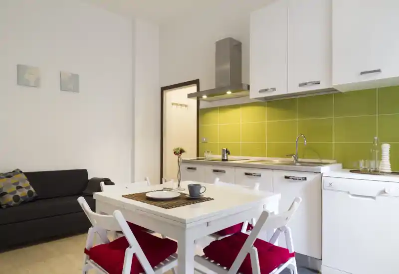 Bright, modern open-plan kitchen with a green tiled backsplash, white cabinets and a small dining table set with red-cushioned chairs — ideal for a compact apartment.