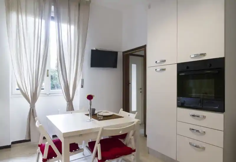 Bright, compact kitchen with a white dining table, red seat cushions, built-in oven and a large window letting in natural light — ideal for a cozy breakfast or small meals.