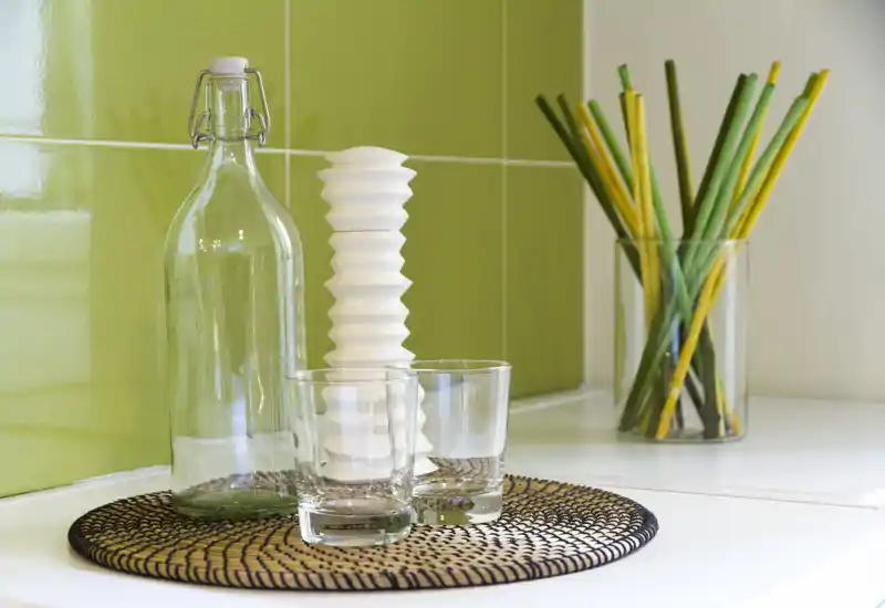 Close-up of a kitchen countertop styled with a glass bottle, two drinking glasses, a decorative white object and a vase with colorful reed sticks against glossy green tiles.