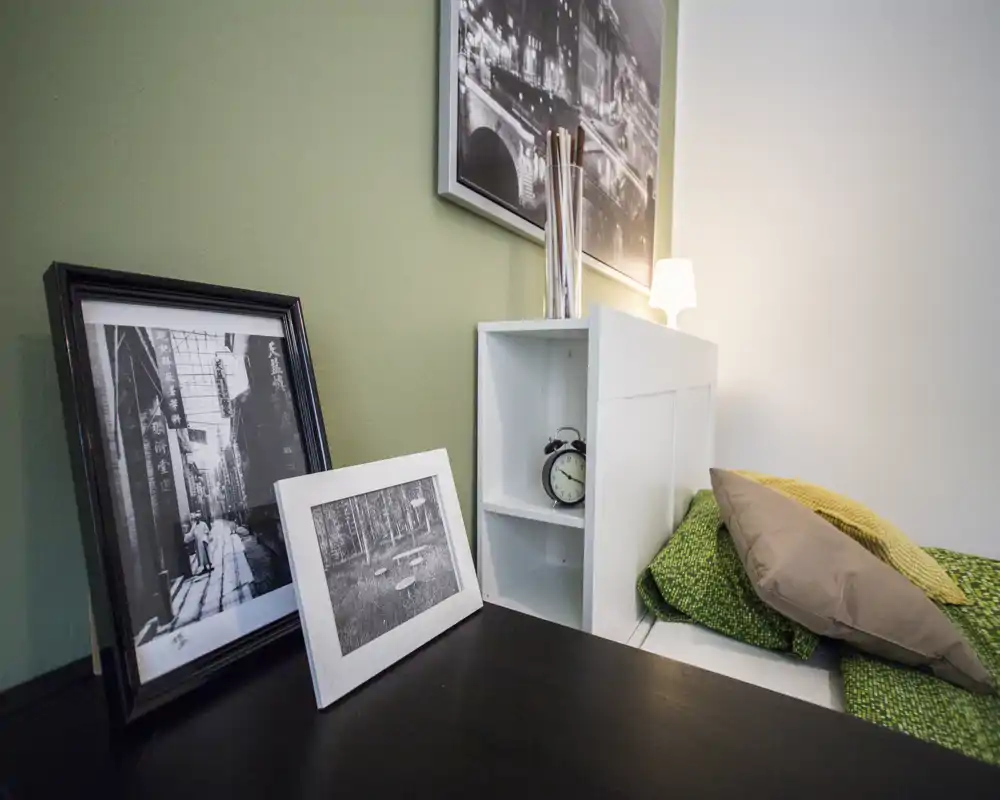 Angolo camera luminoso e accogliente con tessili verdi, cornici decorative e una piccola testiera a scaffale bianca con orologio e lampada.
