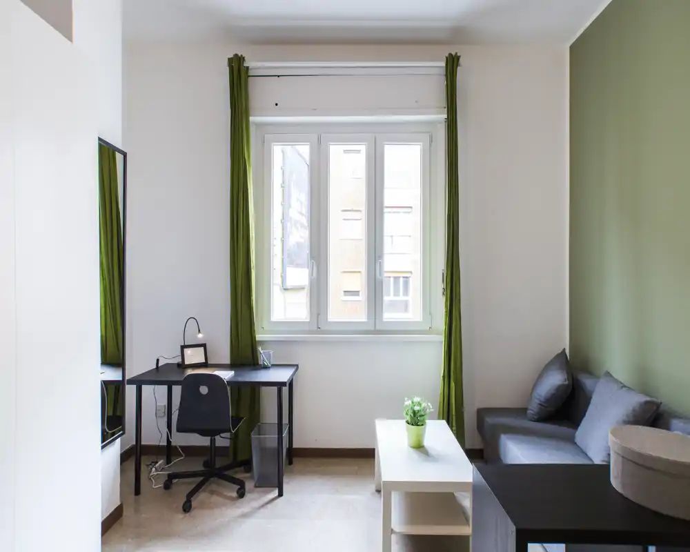 Zona living luminosa e ben allestita con divano compatto, tavolino e scrivania accanto a una finestra ampia; tonalità neutre con accenti verdi creano uno spazio fresco e accogliente, ideale per rilassarsi o lavorare.