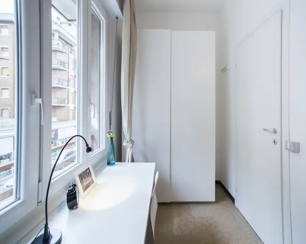 Angolo di camera luminoso con scrivania bianca davanti a grandi finestre e un armadio alto bianco. Luce naturale abbondante; arredo minimale con lampada da tavolo, cornice e un piccolo vaso.