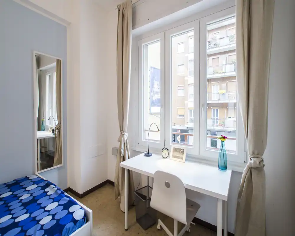 Camera da letto compatta e luminosa con scrivania bianca davanti a una grande finestra, letto semplice con copriletto blu a fantasia, specchio a figura intera e tende chiare — ideale per uno studente o una singola persona.