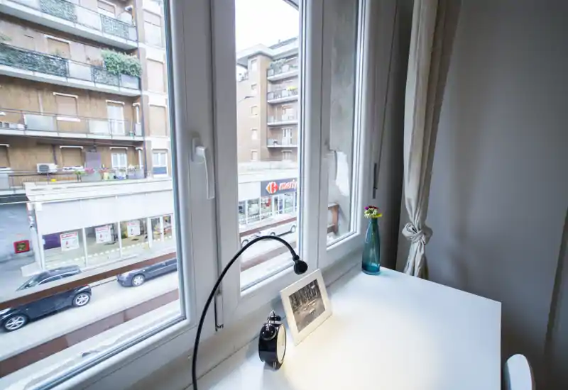 Angolo camera vicino alla finestra con scrivania bianca, lampada, sveglia e vaso decorativo; luce naturale intensa e vista sulla strada dalle ampie finestre.