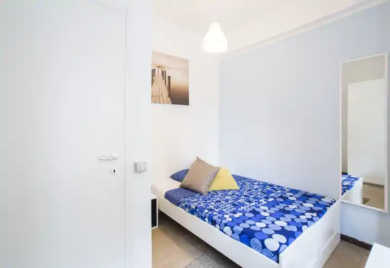 Camera singola compatta con struttura del letto bianca e una vivace biancheria blu a fantasia, arricchita da cuscini neutri e uno specchio a figura intera. La stanza è luminosa, arredata in modo essenziale, ideale per uno studente o un viaggiatore singolo.