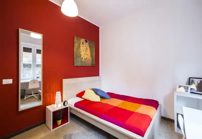 Chambre simple lumineuse et colorée avec un lit bien fait aux couettes rouge-orangé, une table de chevet avec lampe, une décoration murale et une grande glace reflétant la lumière naturelle de la fenêtre.