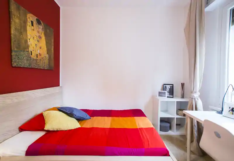 Chambre lumineuse et colorée avec lit double habillé de linge rouge-orangé, coussins décoratifs et petite étagère blanche près de la fenêtre. La lumière naturelle et la décoration minimaliste offrent une ambiance chaleureuse et moderne, idéale pour une annonce locative.