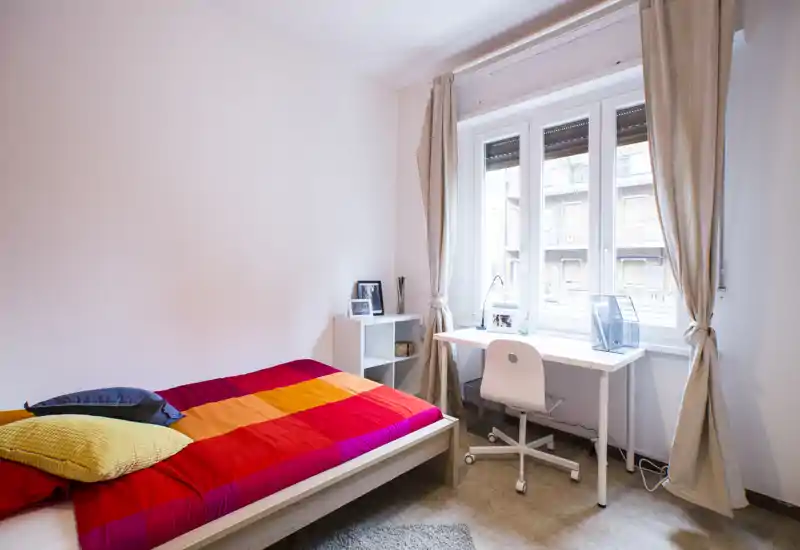 Chambre lumineuse et bien aménagée avec un lit simple coloré, un bureau près d'une grande fenêtre et des rideaux neutres — parfaite pour un étudiant ou une personne seule.