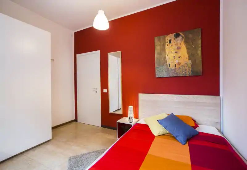 Chambre moderne et lumineuse avec housse de lit colorée, tête de lit, table de chevet et lampe. Le mur rouge et le tableau décoratif apportent une atmosphère chaleureuse et stylée, idéale pour une annonce.