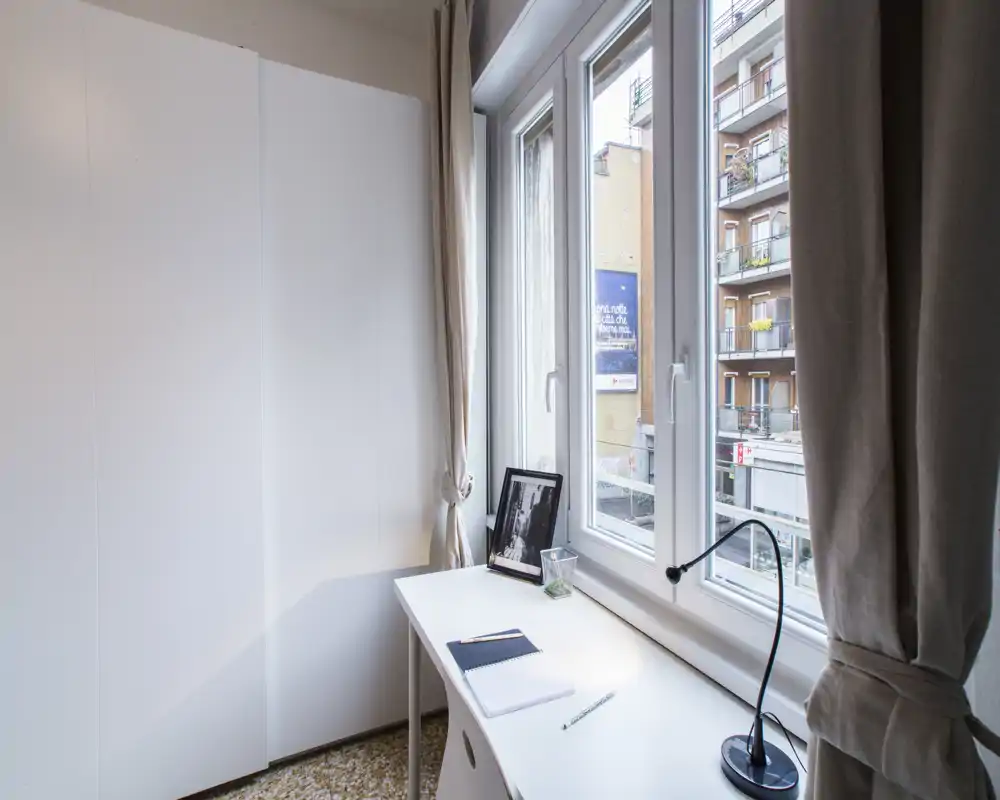 Rincón de dormitorio compacto con armario blanco y escritorio junto a la ventana, luz natural que ilumina el espacio — ideal para una persona o como zona de trabajo en la habitación.