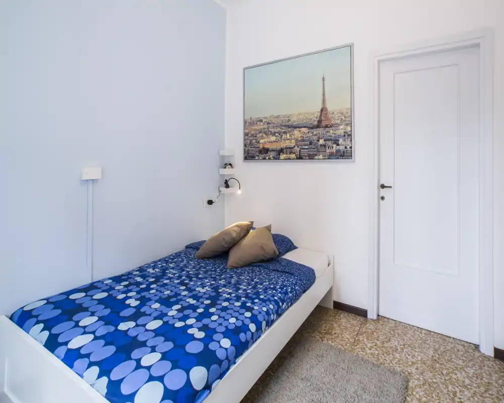 Dormitorio pequeño y luminoso con una cama individual con ropa de cama azul estampada, dos cojines, una lámpara de noche y un cuadro con paisaje urbano sobre la cama. Decoración limpia y sencilla que ofrece un ambiente acogedor para estancias cortas.