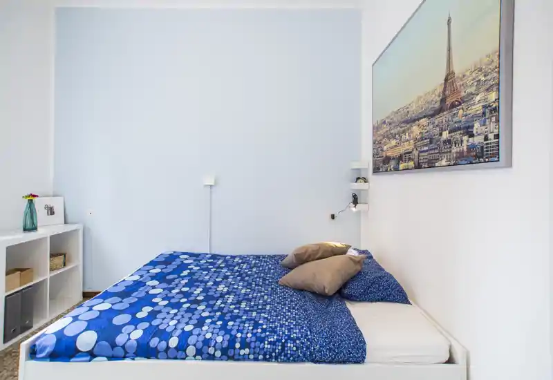 Chambre lumineuse au style épuré avec un lit double recouvert d’une couette bleue à pois, deux oreillers et une grande photo de la ville au-dessus. Étagères propres et décoration minimale pour une ambiance moderne et apaisante, parfaite pour la location courte durée.
