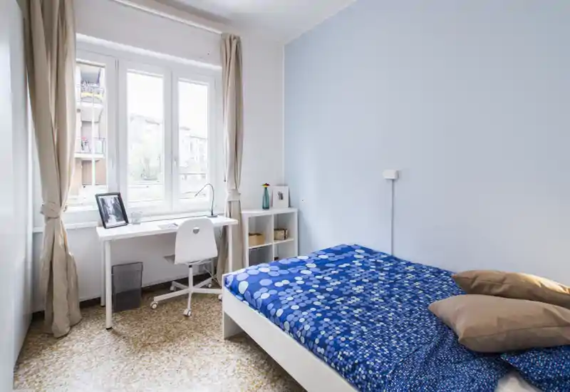 Chambre lumineuse et soignée avec un lit double habillé d'une couette bleue à motifs, un petit bureau près d'une grande fenêtre et un rangement simple — idéale pour une personne ou un couple appréciant un style minimaliste et lumineux.