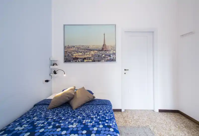Chambre compacte et lumineuse avec une couverture bleue à motifs, deux coussins d'appoint et un cadre représentant une ville au-dessus de la tête de lit. La pièce est sobrement meublée et bien éclairée, idéale pour un court séjour.