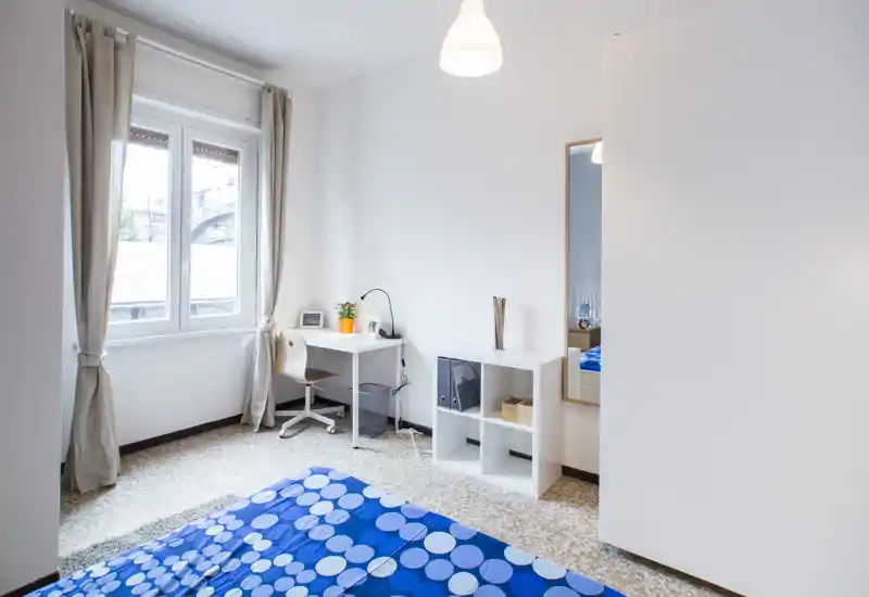 Chambre lumineuse et minimaliste avec une couette bleue à motifs, un bureau près de la fenêtre et des étagères simples. La lumière naturelle, les rideaux neutres et les lignes épurées offrent un espace calme et fonctionnel, parfait pour les courts séjours.