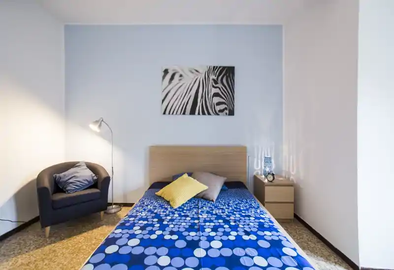 Chambre lumineuse et soignée avec lit double habillé d’une couette bleue à motifs, tête de lit en bois, table de chevet avec lampe et un fauteuil confortable — parfaite pour un séjour agréable.