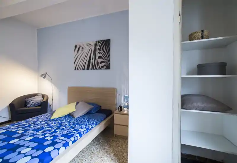 Chambre lumineuse et soignée avec un lit simple habillé d’une couette bleue à motifs, une tête de lit en bois, une table de chevet avec lampe et un fauteuil confortable dans le coin. Décor simple avec une impression zèbre au mur et des étagères intégrées sur le côté.