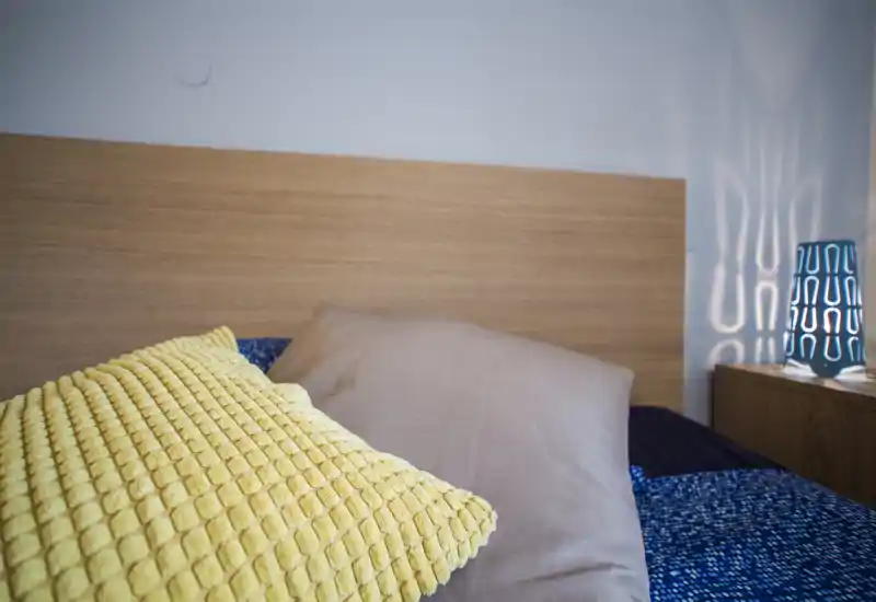 Gros plan d'une chambre lumineuse et confortable avec un coussin jaune texturé, un oreiller beige et une tête de lit en bois; une lampe de chevet diffuse des motifs lumineux.