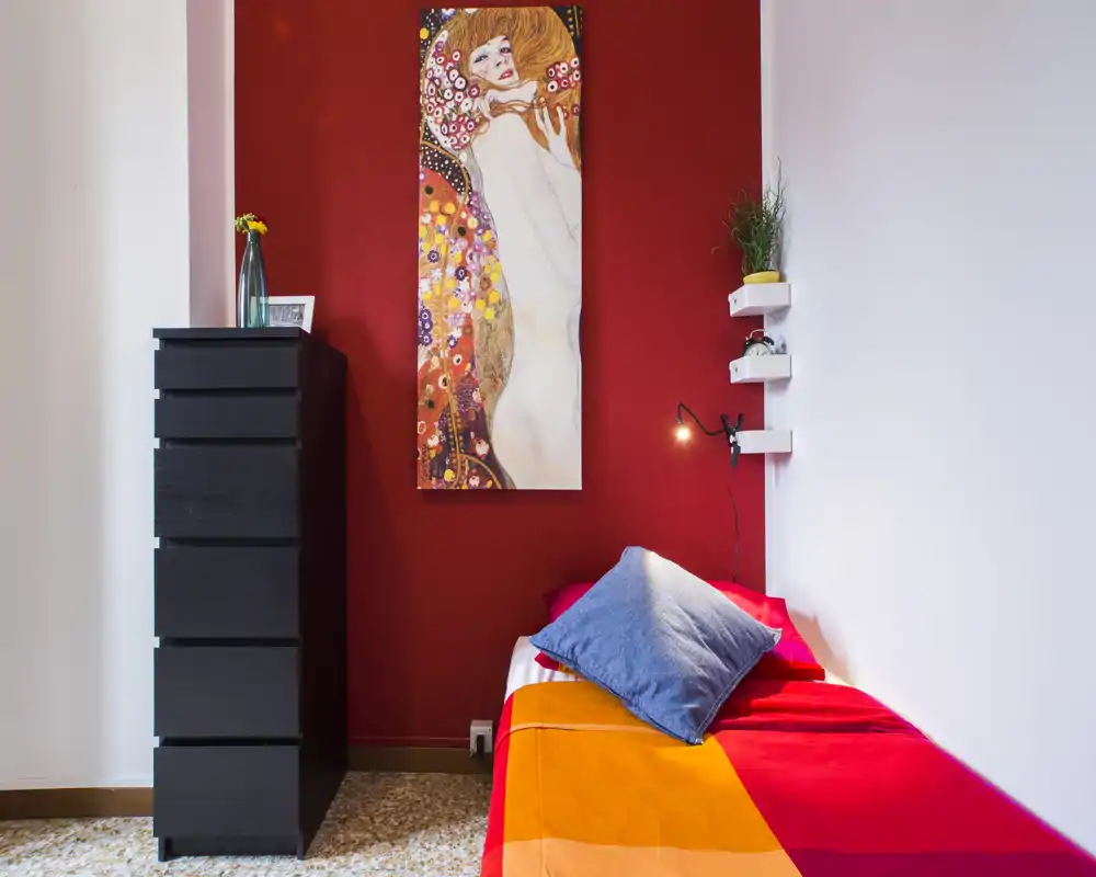 Pequeña habitación colorida con cama individual con ropa de cama brillante, un cojín azul y una pintura vertical llamativa sobre una pared roja. Una alta cómoda negra y estantes flotantes añaden almacenamiento y carácter.