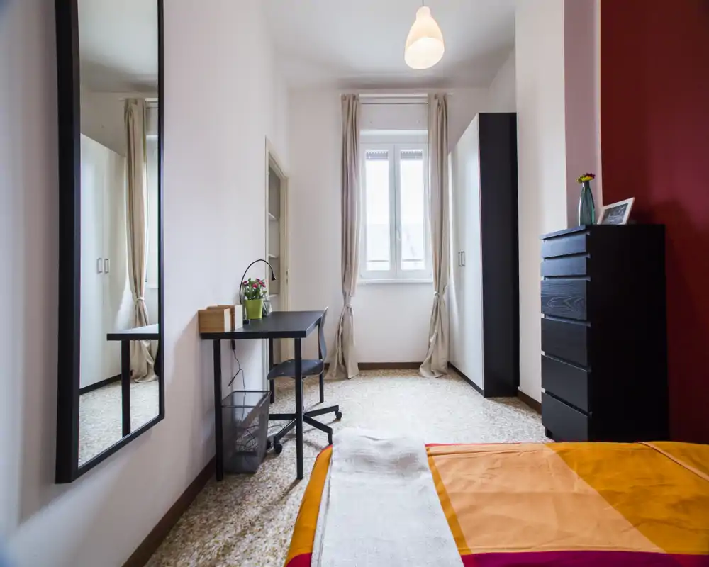 Dormitorio luminoso con cama doble parcialmente visible con ropa de cama naranja, escritorio junto a la ventana, armario y cómoda alta — estancia funcional y bien iluminada, ideal para estancias cortas o largas.