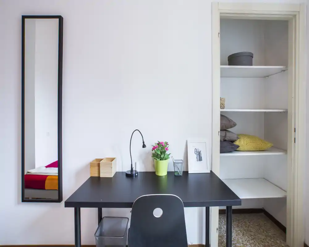 Rincón de dormitorio con mesa negra, silla y un espejo alto que refleja parte de una cama de colores; estantería empotrada con cojines y una planta en la mesa crean un espacio ordenado y luminoso.