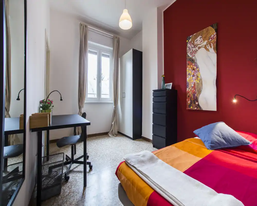 Dormitorio luminoso y acogedor con una cama colorida, lámpara de mesilla y escritorio junto a la ventana. La pared roja y el cuadro crean un ambiente cálido y con estilo, perfecto para un anuncio.