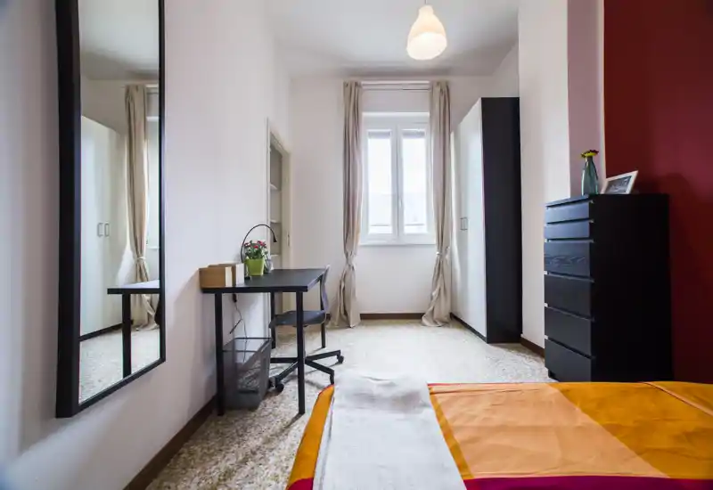 Chambre lumineuse avec lit double partiellement visible et couette orange, bureau près de la fenêtre, armoire et commode haute — pièce fonctionnelle et bien éclairée, parfaite pour courts ou longs séjours.