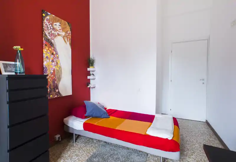 Habitación individual con colcha naranja-roja sobre una cama individual frente a una pared roja de acento. La estancia incluye una cómoda alta negra, un cuadro, una pequeña alfombra y estanterías junto a la cama — luminosa y acogedora para un huésped.